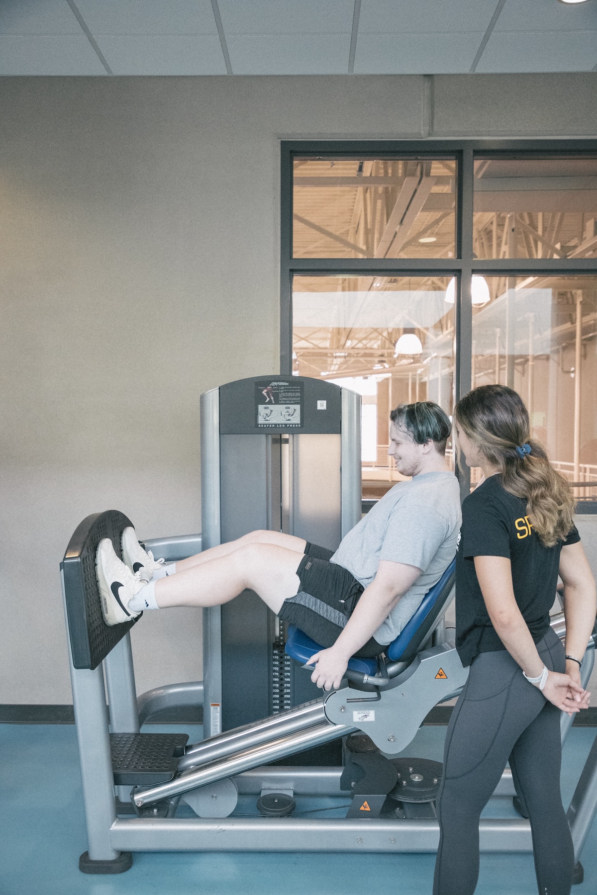 Man exercising on leg machine while a trainer stands nearby